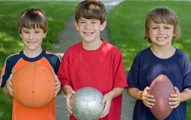 Three boys holding basketball, soccer ball and football